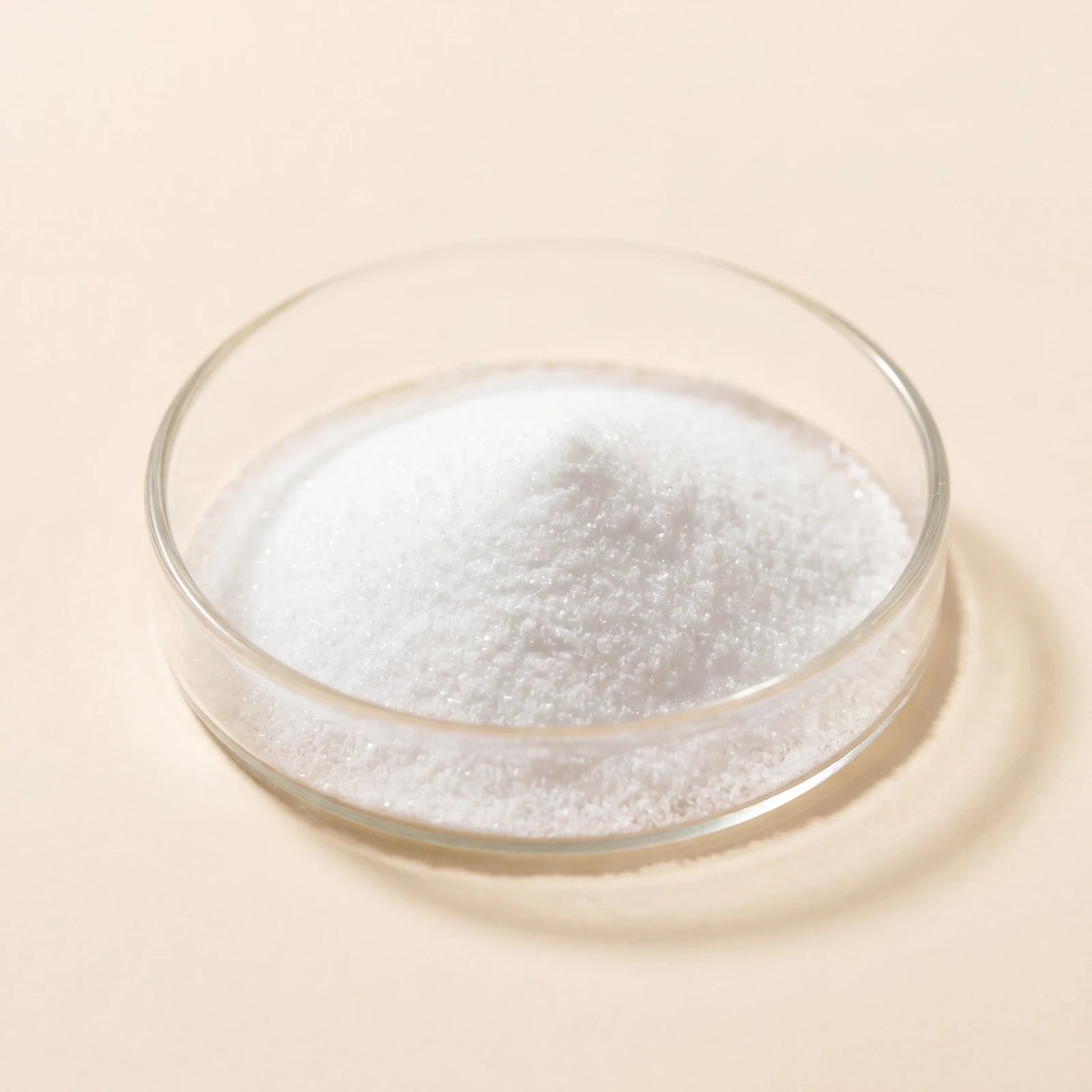 White crystalline niacinamide powder in a glass dish on a cream ground.